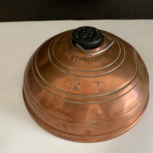 May include: Copper camping lantern with a black lid and a metal handle. The lantern has a circular shape and is embossed with the text "Fill Completely Before Using".