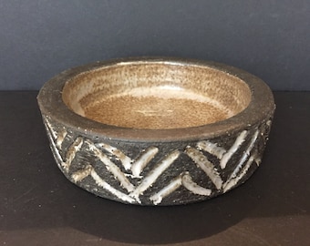 May include: A round ceramic bowl with a brown glaze inside and a black glaze outside. The outside of the bowl has a decorative pattern of white chevron shapes.