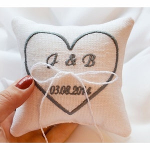May include: A square, off-white ring pillow with a gray embroidered heart design. The heart contains the initials "J & B" and the date "03.08.2014". A white string is tied around the pillow.