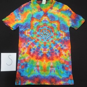 May include: A tie-dye V-neck t-shirt in a vibrant array of colors, including orange, blue, purple, and red. The shirt features a symmetrical spiral pattern radiating from the center. A small white card with the letter "S" is visible.