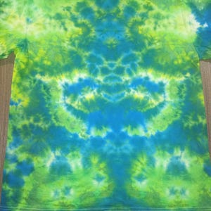 May include: A tie-dye t-shirt in shades of green, yellow, and blue. The shirt has a crew neck and short sleeves. The dye pattern is symmetrical, with a central design and radiating patterns. The shirt is made of a soft, lightweight fabric.