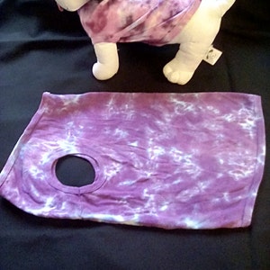May include: Purple and white tie-dye dog shirt with a hole for the head. The shirt is laying flat on a dark purple surface.