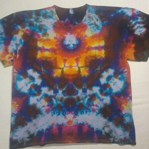 May include: A tie-dye V-neck t-shirt with a colourful pattern. The design features a central orange and yellow burst, surrounded by blue, purple, and brown hues. The shirt is made of soft material.