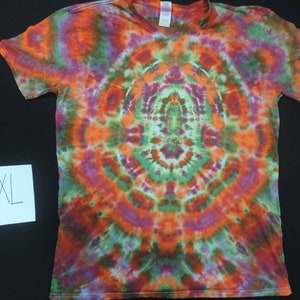 May include: A tie-dye t-shirt with a circular pattern in shades of orange, green, purple, and white. The shirt has a V-neck and short sleeves. The size XL is written on a white piece of paper.