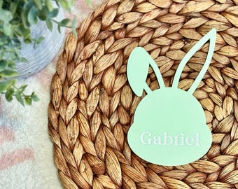 May include: A light green bunny-shaped wooden cutout with the name "Gabriel" written in white on it. The cutout is sitting on a woven brown placemat.