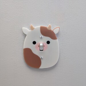 May include: A decorative light switch cover in the shape of a cow's face. The cover is white with brown patches, black eyes, and pink accents. The switch is in the "ON" position.