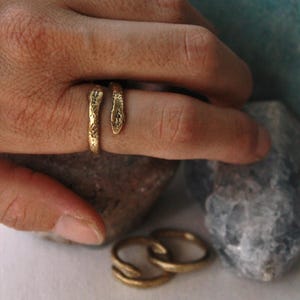 Op de afbeelding: Een goudkleurige metalen ring in de vorm van een kronkelende slang met een getextureerd, onregelmatig oppervlak. De ring heeft twee uiteinden die lijken op een slangenkop en een staart die elkaar niet raken, wat een open ontwerp creëert.