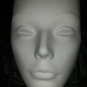 Peut inclure: Un masque en plâtre blanc avec une expression neutre. Le masque a les yeux fermés, un nez légèrement retroussé et une bouche fermée.