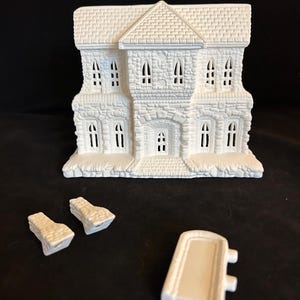 May include: A white ceramic haunted house with a stone-like texture, featuring multiple windows and a front door. The house is accompanied by three detached pieces, likely components for assembly. The overall design suggests a Halloween or holiday decoration.