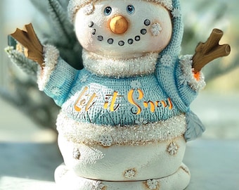 May include: A white snowman figurine wearing a blue knitted hat and sweater. The snowman has a carrot nose, black eyes, and a smiling mouth. The sweater has the words "Let it Snow" written on it. The snowman is standing on a white base.