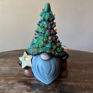 Può includere: Statuetta di gnomo natalizio in ceramica con un cappello a forma di albero verde ornato di luci colorate. Lo gnomo ha una barba grigia, un naso marrone e tiene in mano una stella glitterata. La statuetta è su una superficie di legno.
