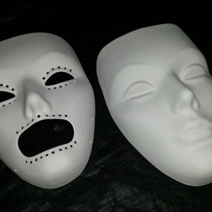 Puede incluir: Dos máscaras de yeso blanco, una con una expresión neutral y la otra con la boca y los ojos muy abiertos, ambas con pequeños agujeros alrededor de los rasgos.