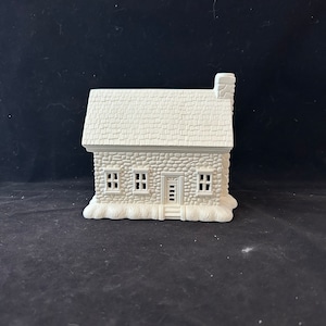 May include: White ceramic house with a stone facade, three windows, and a door. The house has a shingled roof and a chimney. The house is sitting on a base that looks like snow.