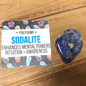 May include: A blue sodalite crystal with white and black flecks. The crystal is on a wooden surface with a card that says "You Found...Sodalite" and "Enhances Mental Powers Intuition - Awareness".