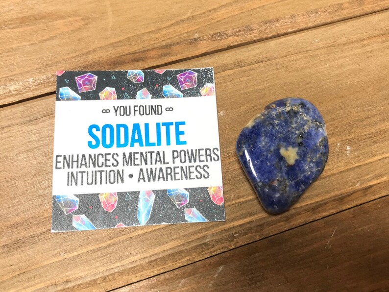 May include: A blue sodalite crystal with white and black flecks. The crystal is on a wooden surface with a card that says "You Found...Sodalite" and "Enhances Mental Powers Intuition - Awareness".