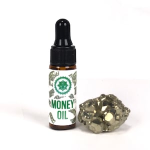 Pode incluir: Um pequeno frasco de vidro castanho com um conta-gotas preto. O frasco é etiquetado como "Money Oil" e tem um design verde e branco com imagens de notas de dólar. Uma pedra de cor dourada está ao lado do frasco.