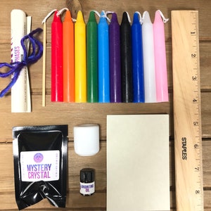 May include: A kit for crystal healing with a mystery crystal, a small bottle of anointing oil, a white candle, a small wooden ruler, a rolled up paper with the text "Moon Phases", a small bag of black powder labeled "Mystery Crystal", and ten small candles in the colors of the rainbow.
