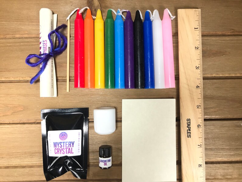 May include: A kit for crystal healing with a mystery crystal, a small bottle of anointing oil, a white candle, a small wooden ruler, a rolled up paper with the text "Moon Phases", a small bag of black powder labeled "Mystery Crystal", and ten small candles in the colors of the rainbow.