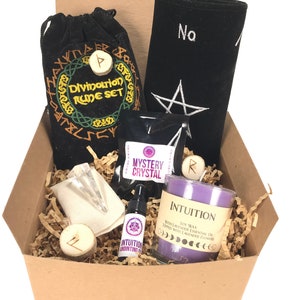 May include: A brown cardboard box filled with a divination rune set, a black cloth with a pentagram, a clear crystal, a small bottle of intuition anointing oil, a purple candle labeled "Intuition", and a mystery crystal. The box is filled with brown packing paper.
