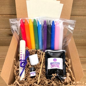 May include: A cardboard box filled with a variety of items for a magical ritual. The box contains a set of rainbow colored candles, a small bottle of anointing oil, a white ceramic dish, a small bag of black crystals labeled "Mystery Crystal", a rolled up piece of paper with the words "Moon Phases" written on it, and a wooden skewer.