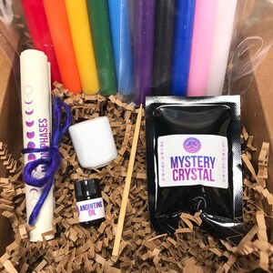 May include: A box filled with brown packing paper contains a set of rainbow colored candles, a small white jar, a small bottle labeled "Anointing Oil", a wooden skewer, a rolled up paper with the text "Moon Phases" and a black pouch labeled "Mystery Crystal".