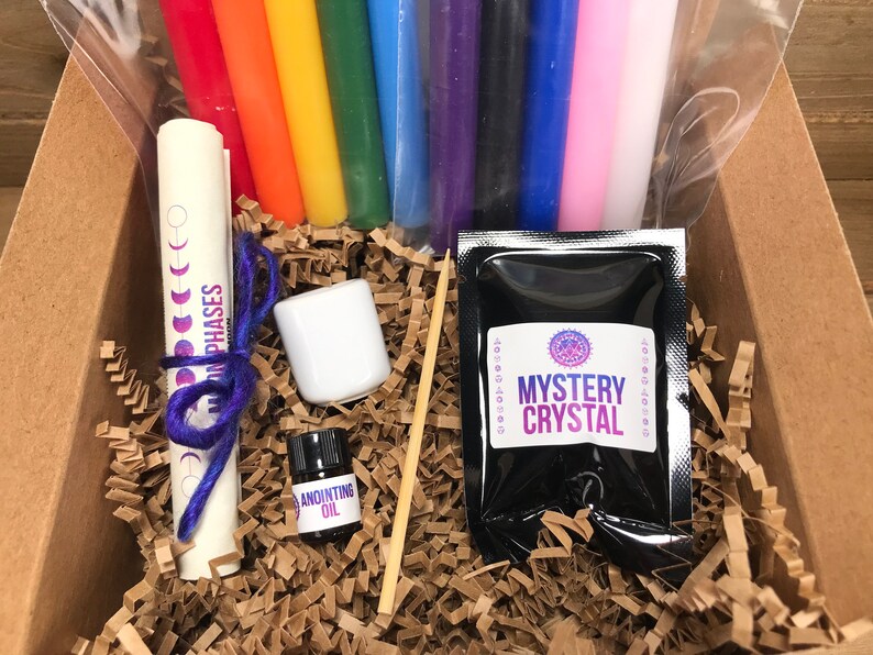 May include: A box filled with brown packing paper contains a set of rainbow colored candles, a small white jar, a small bottle labeled "Anointing Oil", a wooden skewer, a rolled up paper with the text "Moon Phases" and a black pouch labeled "Mystery Crystal".