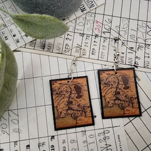 May include: A pair of earrings featuring square-shaped wooden pendants with a vintage map design. The map is printed in brown and black on a beige background. The earrings are attached to silver hooks.