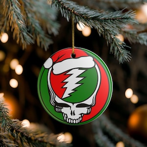 May include: Round Christmas ornament with a red and green design featuring a skull, lightning bolt, and Santa hat. The ornament is hanging from a gold string against a blurred background of Christmas lights and a pine tree.