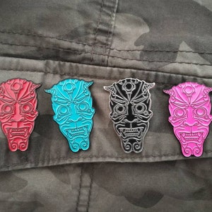 May include: Four enamel pins featuring a stylized Japanese demon mask design. The masks are in red, blue, black, and pink.