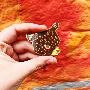 May include: A brown and black enamel pin featuring a cartoon bear face. The bear has a yellow nose and black eyes.