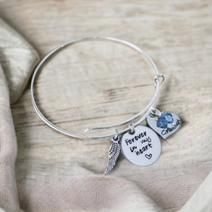 May include: Silver bangle bracelet with three charms. One charm is a wing, another is a heart with a blue crystal, and the third is a round charm with the words "Forever in my heart" and a small heart symbol.