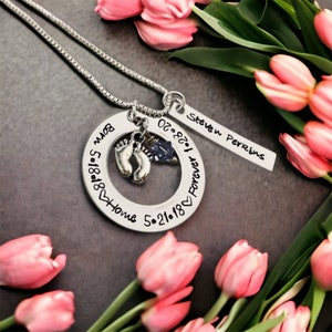May include: Silver necklace with a round pendant that says "Forever Love 02.28.2018" and "Home 5.21.2018 Born 5.18.2018". The pendant has a small charm of two baby footprints and a blue crystal. A silver tag with the text "Steven Perrins" hangs from the pendant.