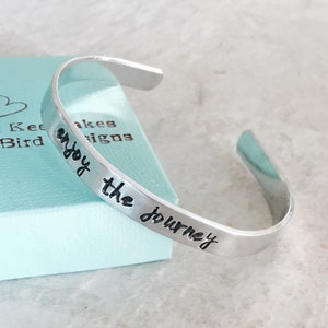 May include: Silver cuff bracelet with the words "enjoy the journey" engraved on it.