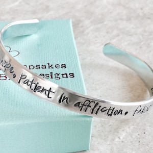 Puede incluir: Pulsera de plata con la inscripción "Sé paciente en la aflicción, la fe es la esperanza." La pulsera está sobre una caja azul claro con las palabras "Crafted by Shirley B. Designs" en ella.