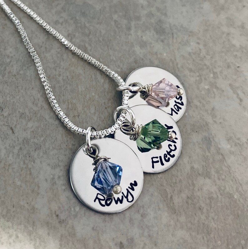 May include: A silver chain necklace with three silver disc charms. Each charm has a different colored crystal and a name engraved on it: Rowyn, Fletcher, and Mais.