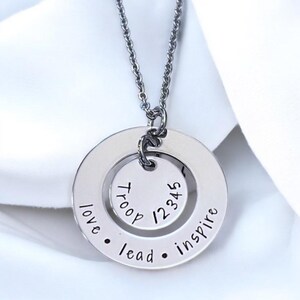Könnte beinhalten: Eine silberfarbene Halskette mit Anhänger und zwei kreisförmigen Charms. Der äußere Charm trägt die eingravierten Worte "love • lead • inspire". Der innere Charm zeigt das Wort "Troop" und die Zahlen "12345".