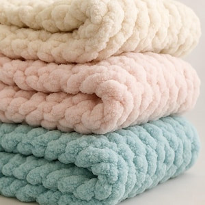 Può includere: Una pila di tre coperte a maglia grossa in morbidi colori pastello. La coperta superiore è bianco crema, seguita da un rosa pallido, e quella inferiore è azzurra. Il filato spesso crea un aspetto accogliente e strutturato.