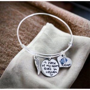 May include: Silver charm bracelet with a heart charm that says "Oliver" and a round charm that says "A piece of my heart lives in Heaven". The bracelet also has a small angel wing charm.