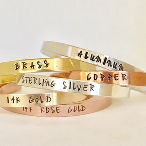 May include: A stack of five different metal bracelets, each with a different metal type engraved on them. The bracelets are stacked in order from top to bottom: Aluminum, Brass, Sterling Silver, Copper, 14k Gold, and 14k Rose Gold.