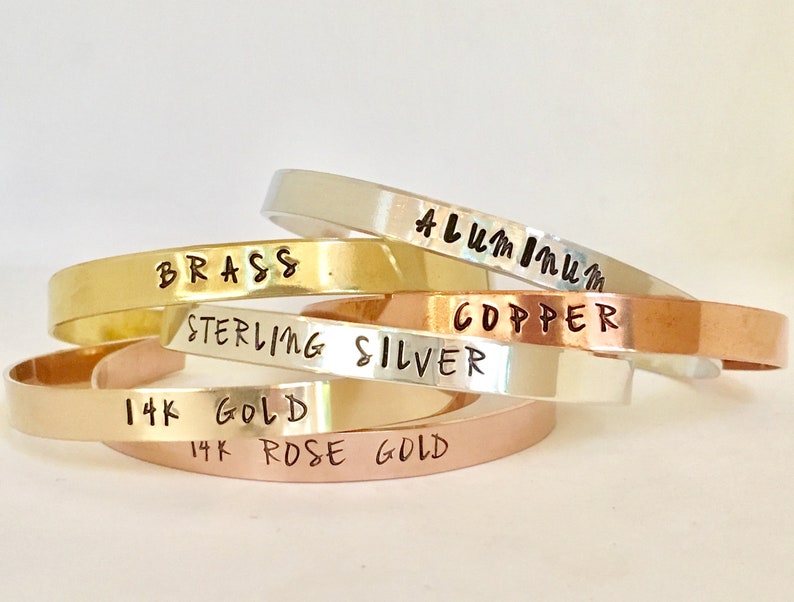 May include: A stack of five different metal bracelets, each with a different metal type engraved on them. The bracelets are stacked in order from top to bottom: Aluminum, Brass, Sterling Silver, Copper, 14k Gold, and 14k Rose Gold.