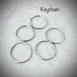 Puede incluir: Cinco llaveros plateados sobre una superficie gris. La palabra "Keychain" est&aacute; escrita sobre los llaveros.