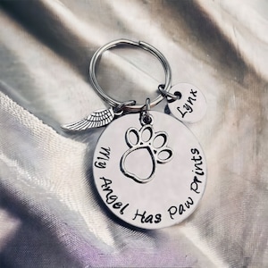 May include: Silver keychain with a round charm that says "My Angel Has Paw Prints" with a paw print cutout. The keychain also has a small angel wing charm and a round charm that says "Lynx".