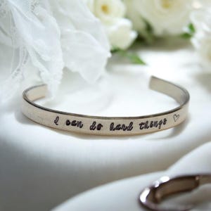 Könnte beinhalten: Silbernes Armreif-Armband mit der Inschrift "Ich kann schwere Dinge tun" und einem kleinen Herzsymbol.