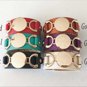 May include: A stack of five leather bracelets with gold-toned metal accents and a round, engraved plate. The bracelets are in the colors red, teal, black, orange, and white.