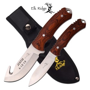 May include: A set of two hunting knives with wooden handles and a black nylon sheath. The knives have a stainless steel blade and a hook on the end of one blade. The sheath has the text "Elk Ridge" embroidered on it. The knife with the hook has the text "JUSTIN 4-12-19" engraved on the blade.