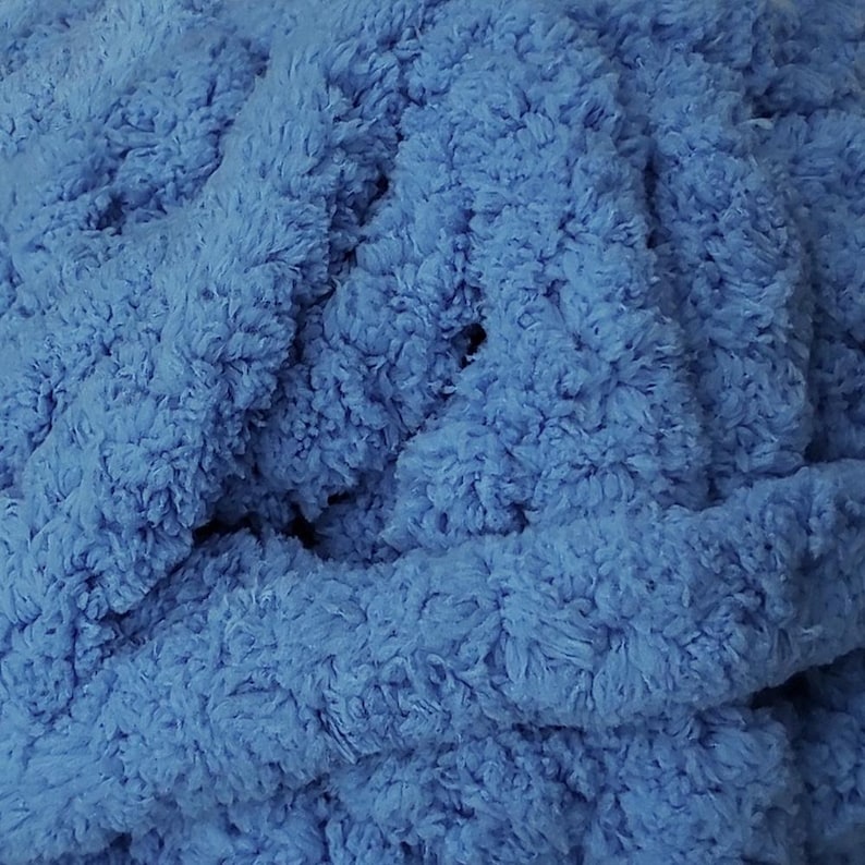 May include: Close-up of a soft, textured, blue fabric. The material has a plush, fluffy appearance, with visible fibers creating a dense, tactile surface. The color is a vibrant, medium blue, filling the frame with its cozy texture.