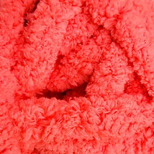 May include: Close-up of a vibrant coral-colored, fluffy textile. The texture appears soft and plush, with a dense, irregular surface. The material is tightly woven, creating a thick, tactile appearance.