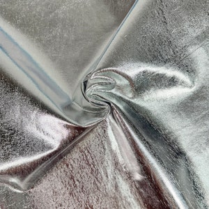 Puede incluir: Tela met&aacute;lica plateada con un patr&oacute;n tejido y texturizado. La tela est&aacute; doblada y drapeada, mostrando su brillo y textura.