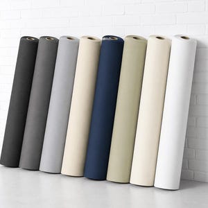 May include: Eight fabric rolls in a range of colours, from dark grey to cream and navy, are propped against a white brick wall. The rolls display a variety of shades and textures, suitable for different design projects.