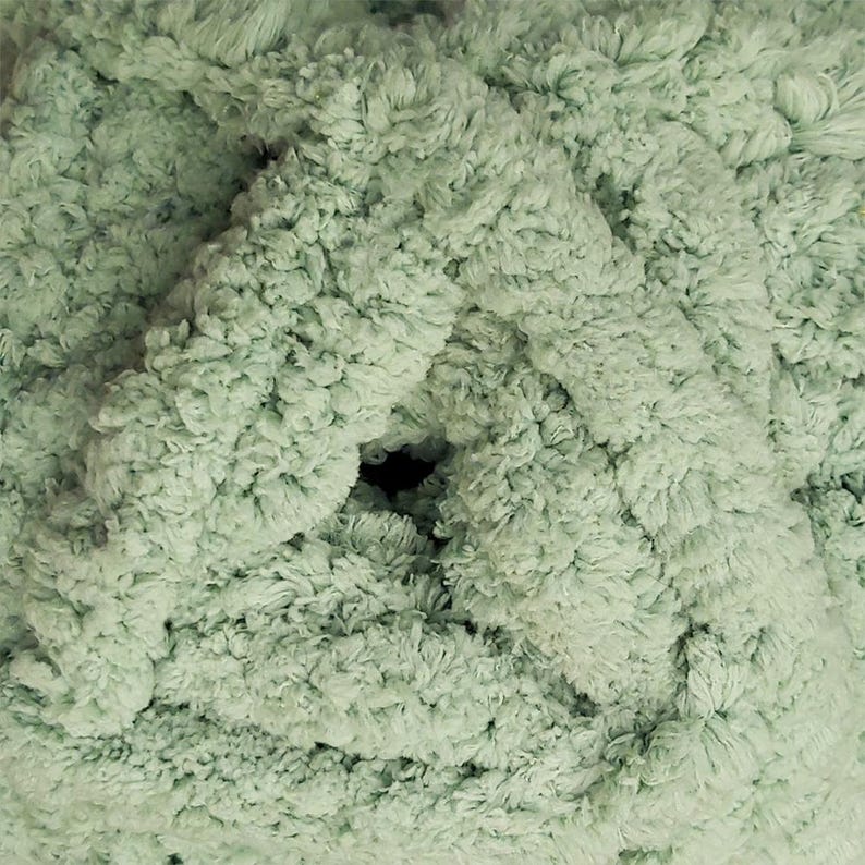 May include: Close-up of a ball of soft, textured yarn in a light green color. The yarn has a fluffy, looped texture, creating a dense and plush appearance. The center of the ball has a dark hole. Ideal for knitting and crochet projects.